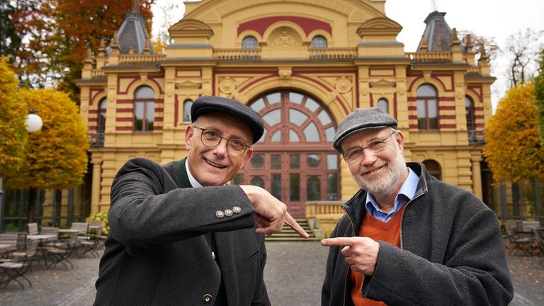 Thomas Schwartz und Harald Lesch stehen vor einem Theater. Beide schauen lächelnd in die Kamera. Jeder zeigt auf den jeweils anderen.