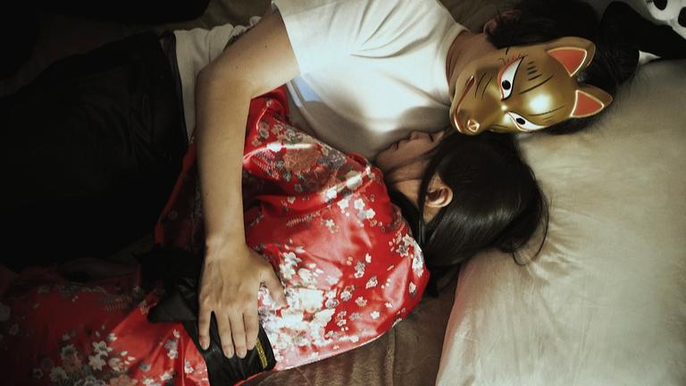 Ein Mann mit goldener Maske und eine Frau im roten Kimono liegen eng umschlungen im Bett. Sie drückt ihr Gesicht an seine Brust. 