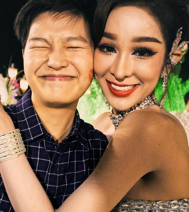 Frontalaufnahme eines jungen Mannes im Karohemd und einer festlich gekleideten Frau mit auffälligem Silberschmuck. Sie legt ihm lachend die Arme um den Oberkörper und er kneift die Augen zu.