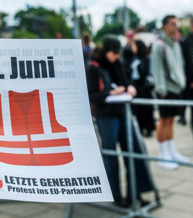 Eine Hand hält einen Flyer der Letzten Generation für die Wahl zum EU-Parlament am 9. Juni. Im Hintergrund sind viele Menschen verschwommen zu erkennen.