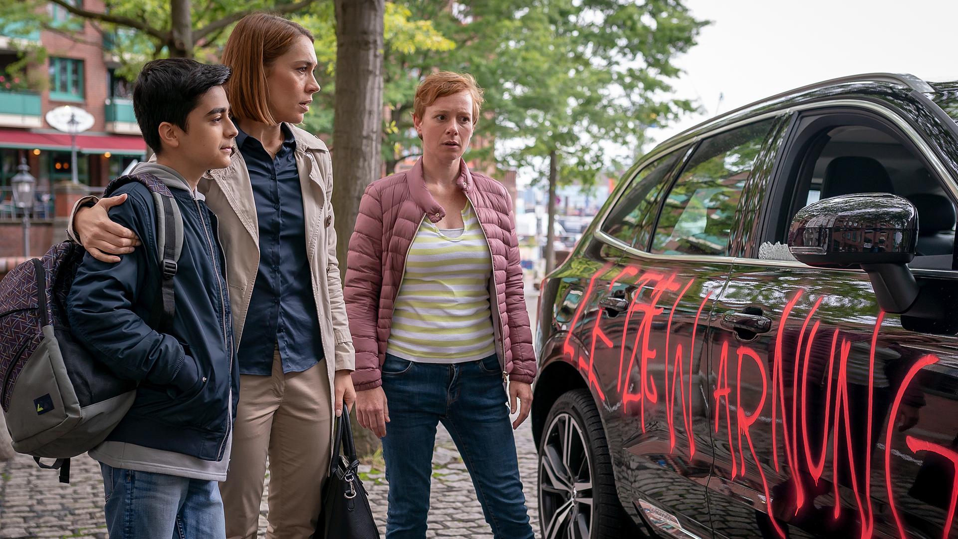Berat (Ali-Emre Sahin), Anne (Henrike Hahn) und Katja (Caroline Junghanns) stehen nebeneinander auf einem Gehweg aus Kopfsteinpflaster. Anne hat ihren Arm um Berats Schulter gelegt. Alle schauen auf die Beifahrerfront eines schwarzen PKW, auf der in roter Schrift "Letzte Warnung" steht.