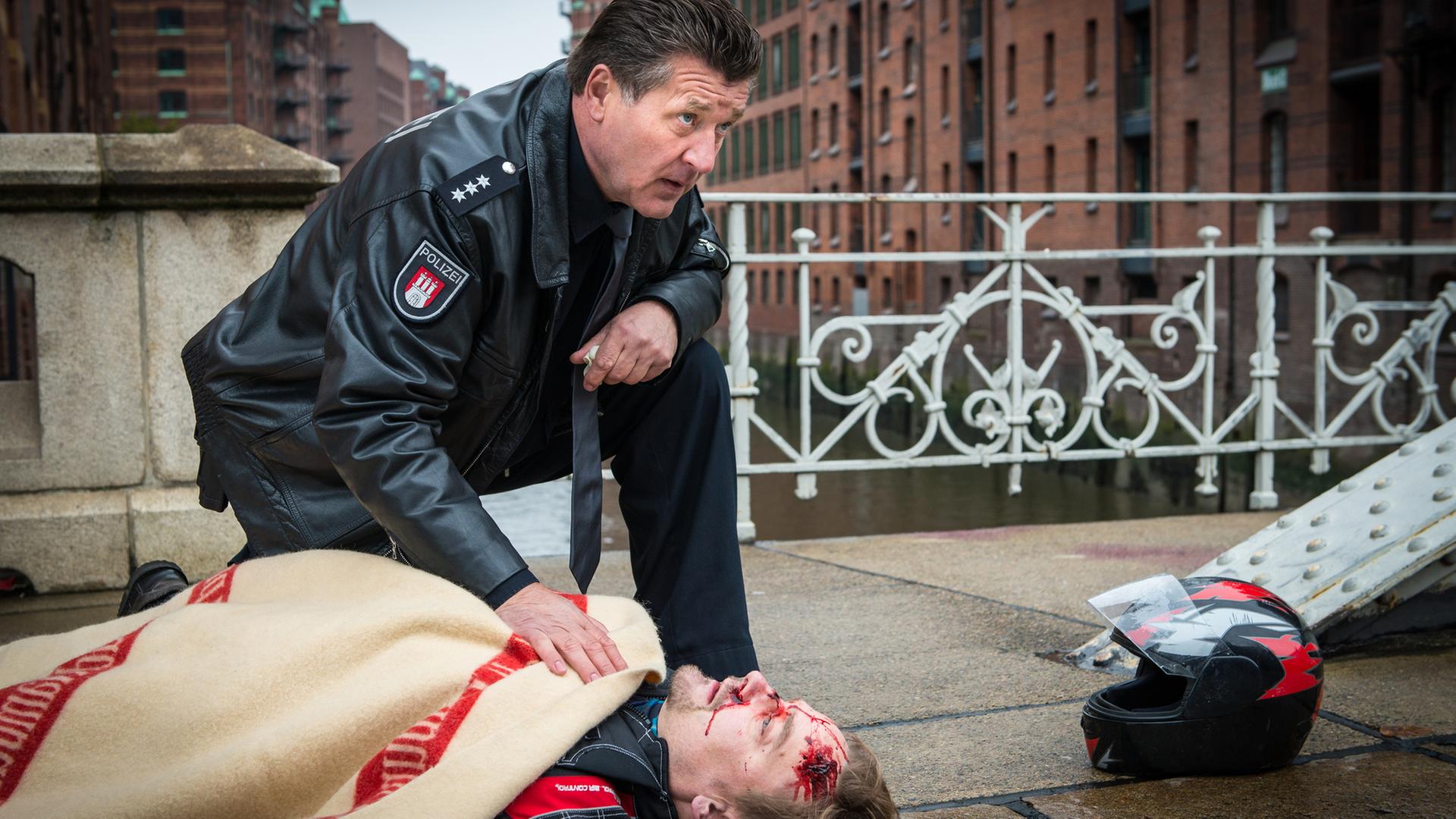 "Notruf Hafenkante - Letzte Worte": Der Polizeibeamte Hans Moor (Bruno F. Apitz) kniet neben dem verletzten Thies Amman (Christian Beermann), der auf dem Bürgersteig auf dem Rücken liegt und mit einer Decke bedeckt ist. Neben ihm liegt ein Motorradhelm. Im Hintergrund: Gebäude der Speicherstadt.