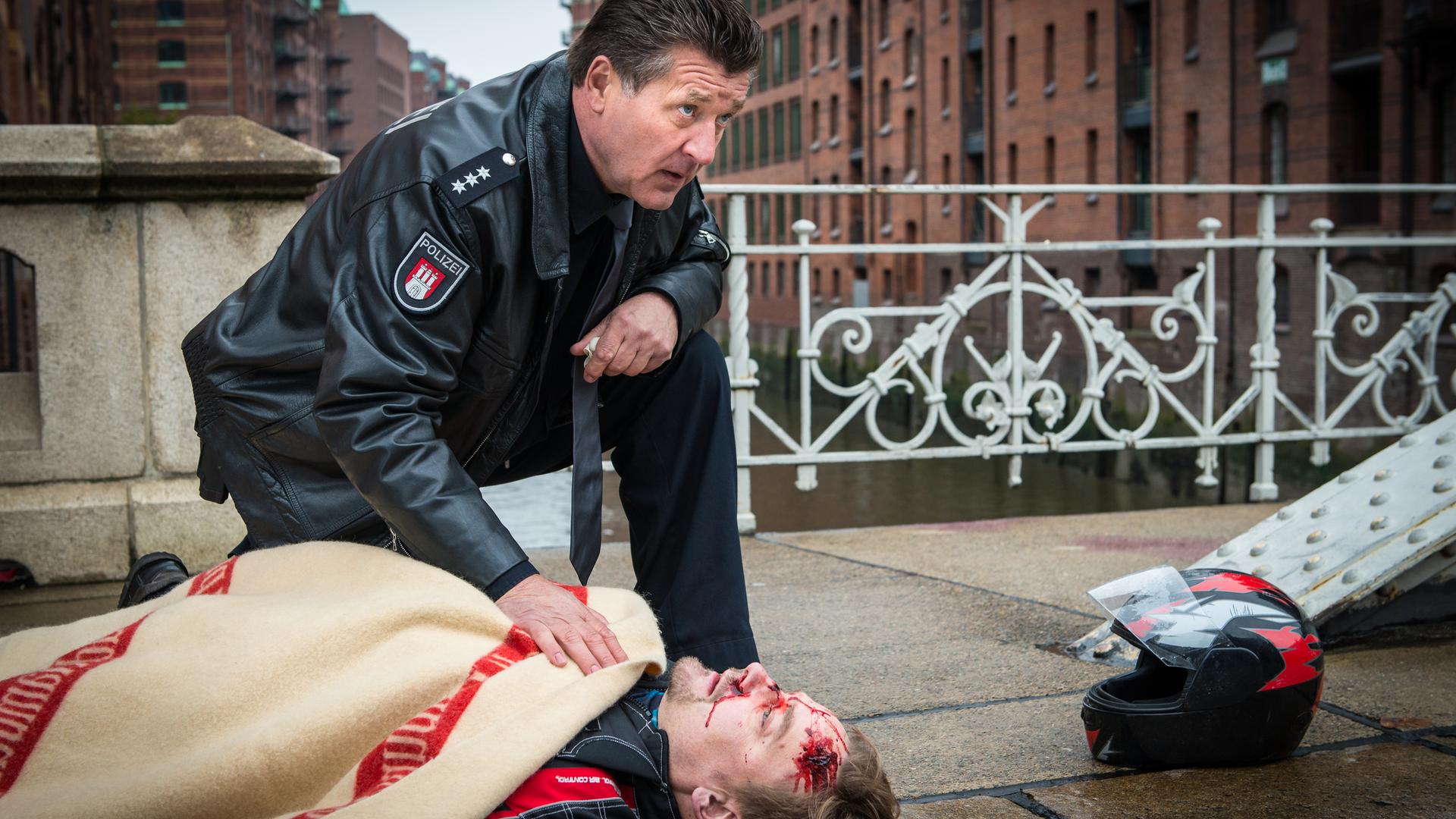 "Notruf Hafenkante - Letzte Worte": Der Polizeibeamte Hans Moor (Bruno F. Apitz) kniet neben dem verletzten Thies Amman (Christian Beermann), der auf dem Bürgersteig auf dem Rücken liegt und mit einer Decke bedeckt ist. Neben ihm liegt ein Motorradhelm. Im Hintergrund: Gebäude der Speicherstadt.