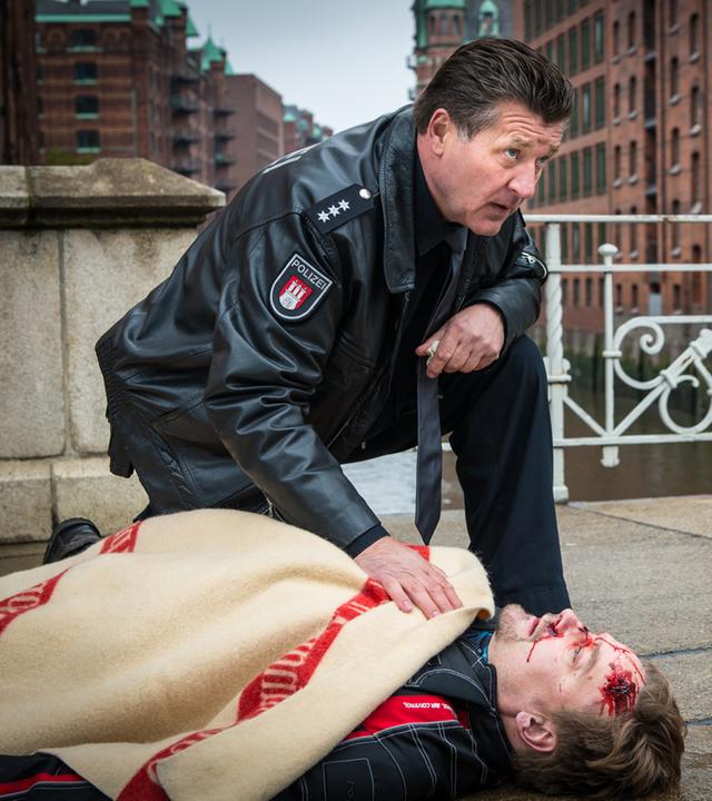 "Notruf Hafenkante - Letzte Worte": Der Polizeibeamte Hans Moor (Bruno F. Apitz) kniet neben dem verletzten Thies Amman (Christian Beermann), der auf dem Bürgersteig auf dem Rücken liegt und mit einer Decke bedeckt ist. Neben ihm liegt ein Motorradhelm. Im Hintergrund: Gebäude der Speicherstadt.