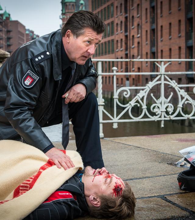 "Notruf Hafenkante - Letzte Worte": Der Polizeibeamte Hans Moor (Bruno F. Apitz) kniet neben dem verletzten Thies Amman (Christian Beermann), der auf dem Bürgersteig auf dem Rücken liegt und mit einer Decke bedeckt ist. Neben ihm liegt ein Motorradhelm. Im Hintergrund: Gebäude der Speicherstadt.