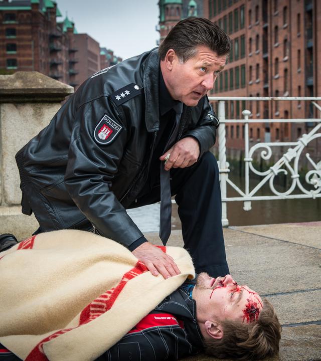 "Notruf Hafenkante - Letzte Worte": Der Polizeibeamte Hans Moor (Bruno F. Apitz) kniet neben dem verletzten Thies Amman (Christian Beermann), der auf dem Bürgersteig auf dem Rücken liegt und mit einer Decke bedeckt ist. Neben ihm liegt ein Motorradhelm. Im Hintergrund: Gebäude der Speicherstadt.