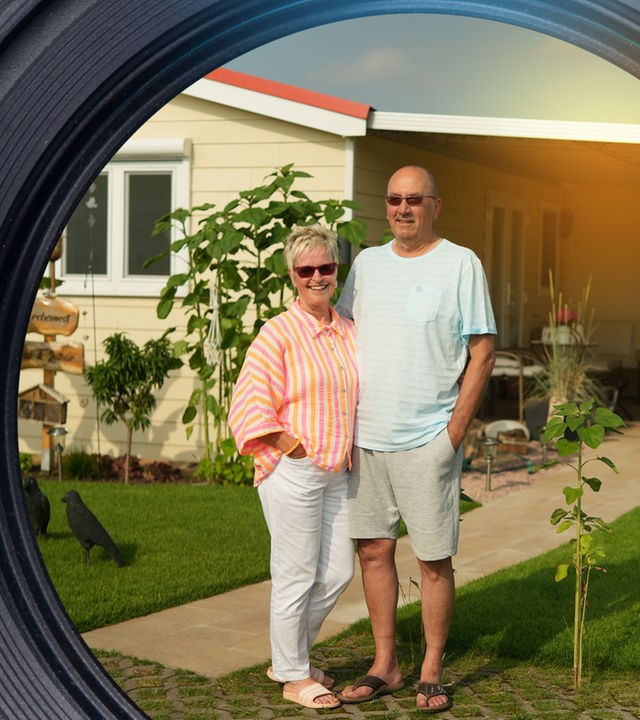 Beatrix und Lothar stehen vor dem Grundstück eines Mobilheims. Beide sind etwa Anfang 60 und schauen in die Kamera. Hinter dem Paar auf der Wiese sitzen drei Rabenstatuen. Vor dem beige-gelben Mobilheim mit rotem Dach stehen Bananenpflanzen und Sonnenblumen. Außerdem ein Pfahl mit zwei Holzschildern und einem Insektenhotel.
