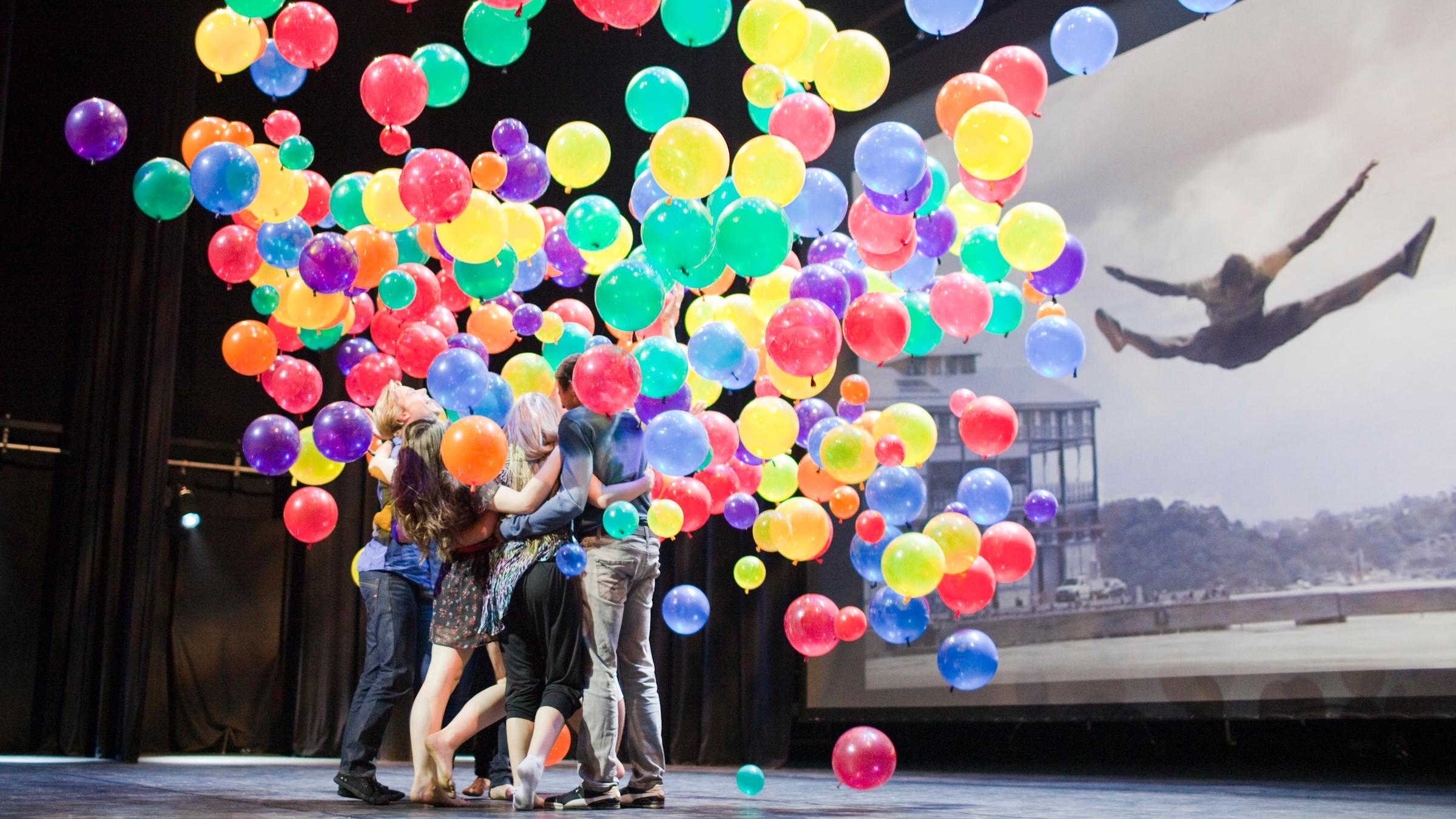 Alle Tänzer umarmen sich und halten ganz viele bunte Luftballons. 