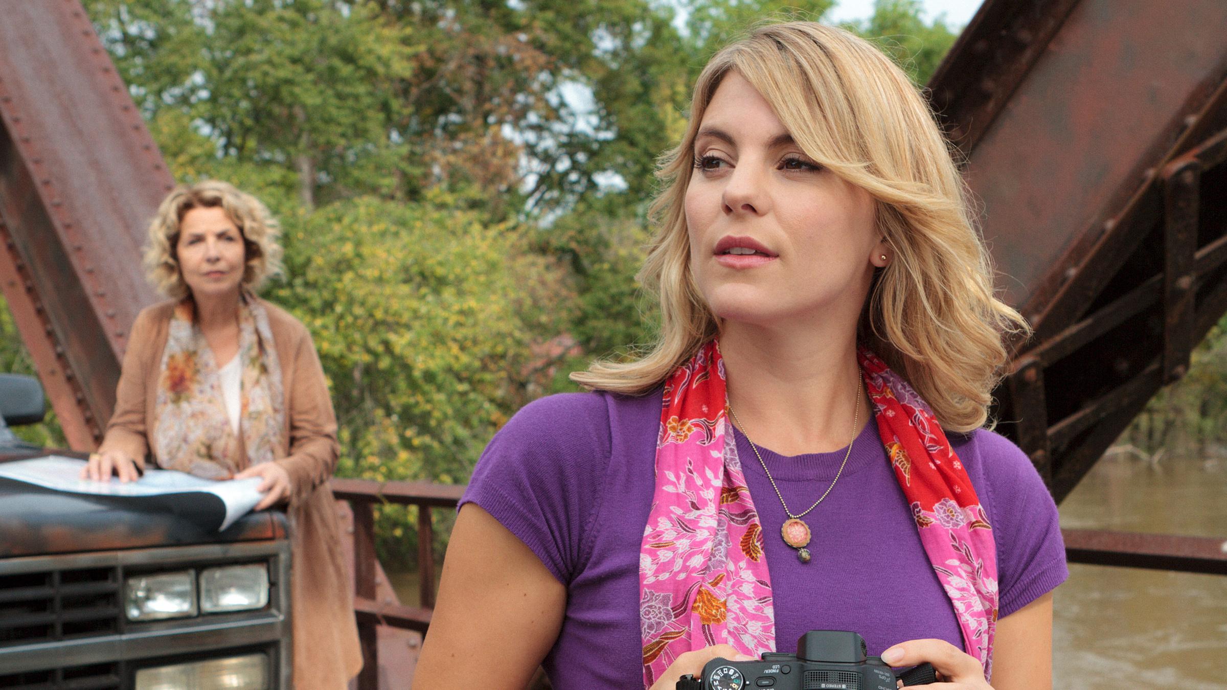 Mona Sadler (Michaela May) und Anna Wilcox (Luise Bähr) stehen mit ihrem Auto auf einer Brücke. Anna hat eine Kamera in der Hand.