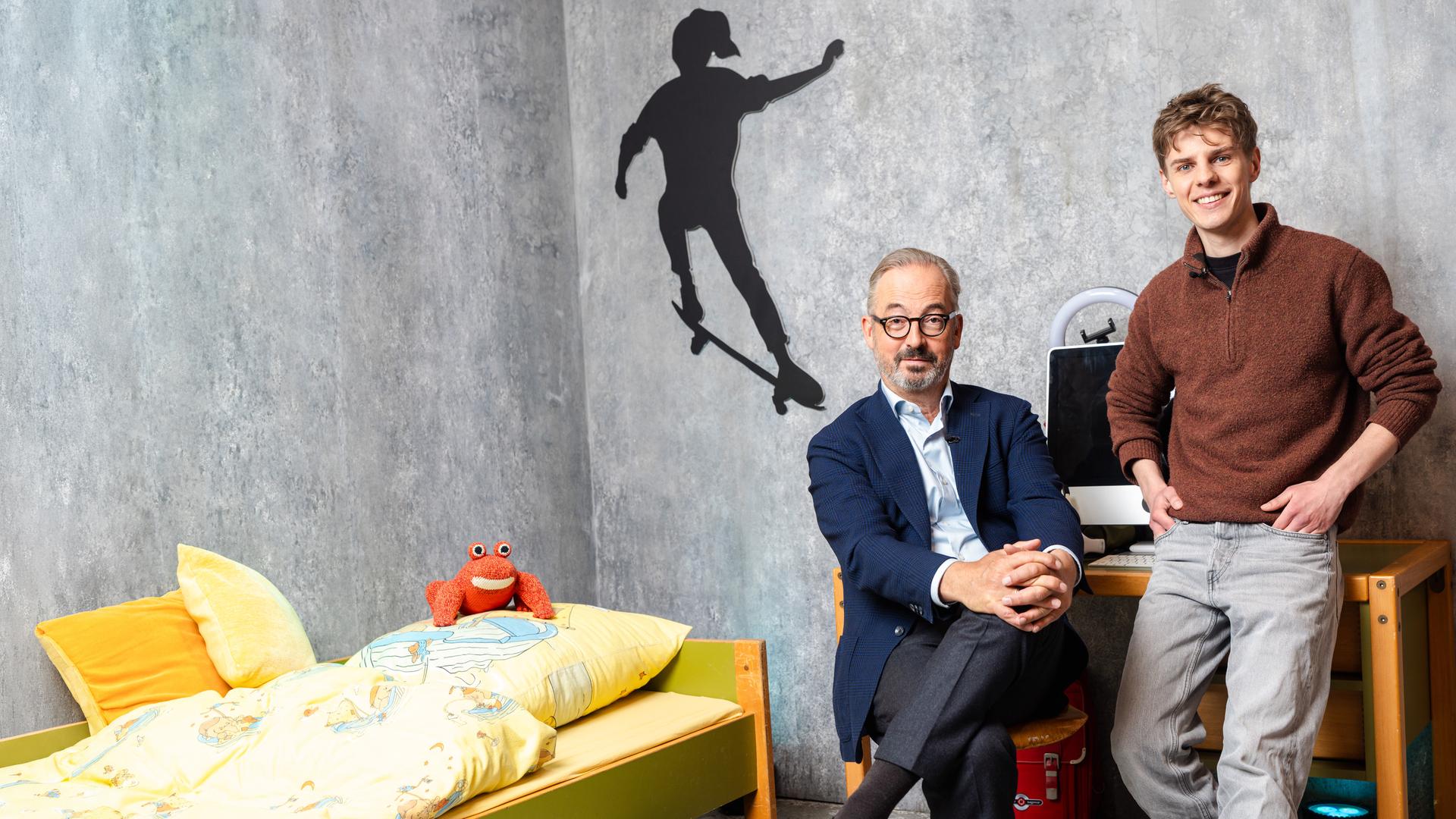 Das Bild zeigt Jan Fleischhauer und Levi Penell in einem Raum mit grauer Betonwand, der wie ein Kinder- oder Jugendzimmer inszeniert ist. Links im Bild steht ein niedriges Bett mit gelber Bettwäsche und bunten Motiven, darauf liegt ein kleines rotes Kuscheltier. An der Wand darüber ist die schwarze Silhouette eines Skateboarders zu sehen.