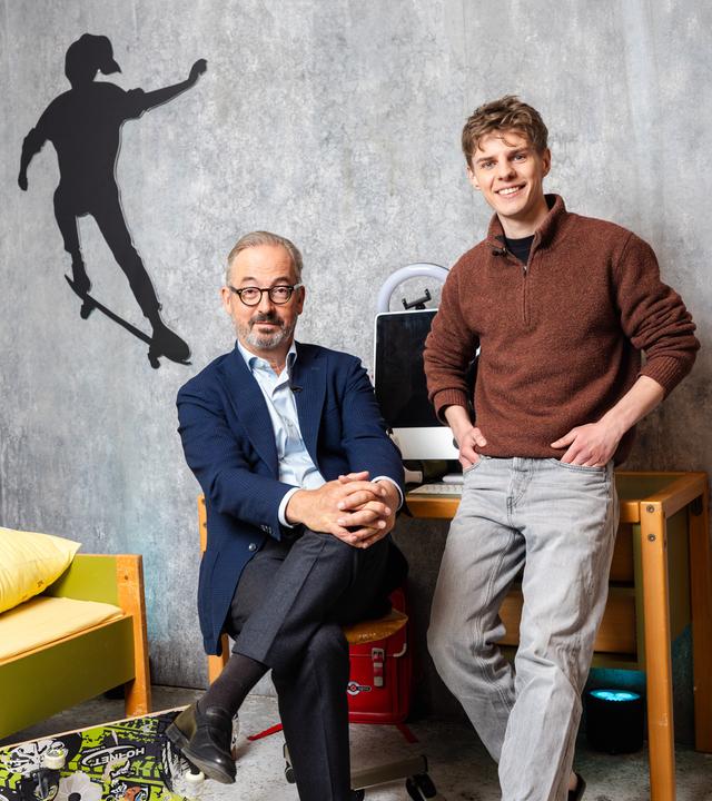 Das Bild zeigt Jan Fleischhauer und Levi Penell in einem Raum mit grauer Betonwand, der wie ein Kinder- oder Jugendzimmer inszeniert ist. Links im Bild steht ein niedriges Bett mit gelber Bettwäsche und bunten Motiven, darauf liegt ein kleines rotes Kuscheltier. An der Wand darüber ist die schwarze Silhouette eines Skateboarders zu sehen.