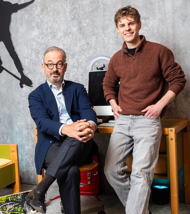 Das Bild zeigt Jan Fleischhauer und Levi Penell in einem Raum mit grauer Betonwand, der wie ein Kinder- oder Jugendzimmer inszeniert ist. Links im Bild steht ein niedriges Bett mit gelber Bettwäsche und bunten Motiven, darauf liegt ein kleines rotes Kuscheltier. An der Wand darüber ist die schwarze Silhouette eines Skateboarders zu sehen.