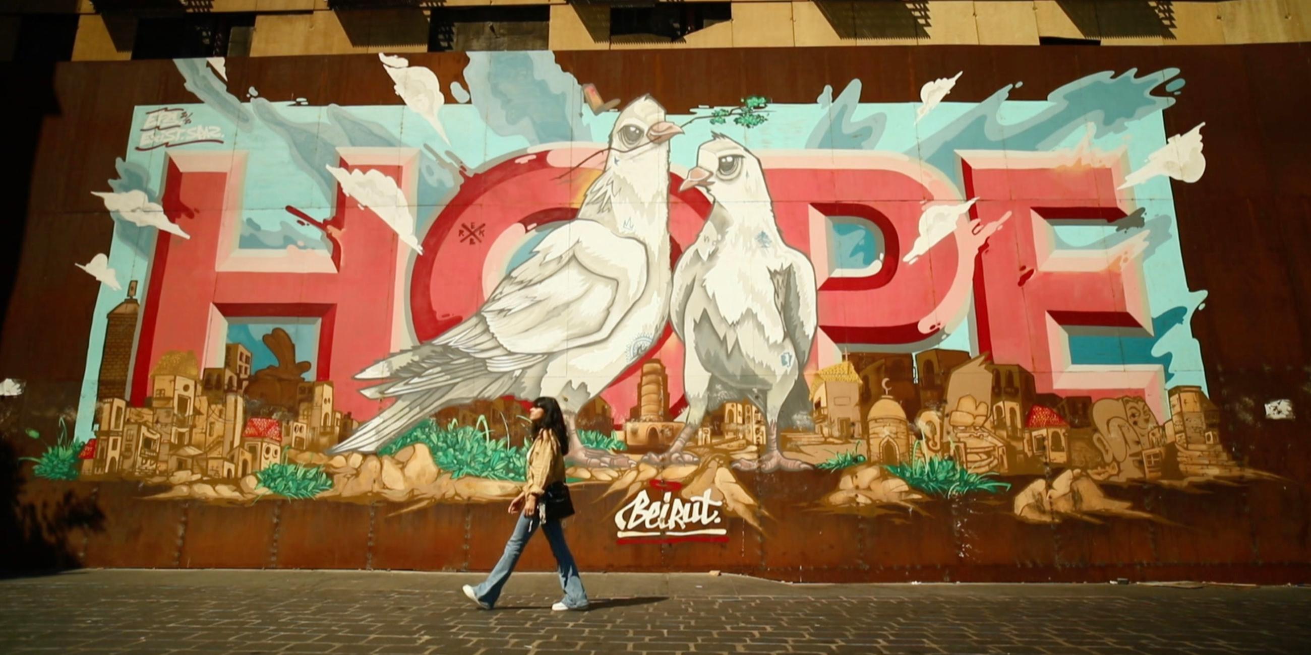  Auf eine Mauer sind zwei große weiße Tauben gemalt, die in einer Stadt stehen. Hinter ihnen ist das Wort "Hope" zu lesen. Vor der Mauer läuft eine junge Frau vorbei.