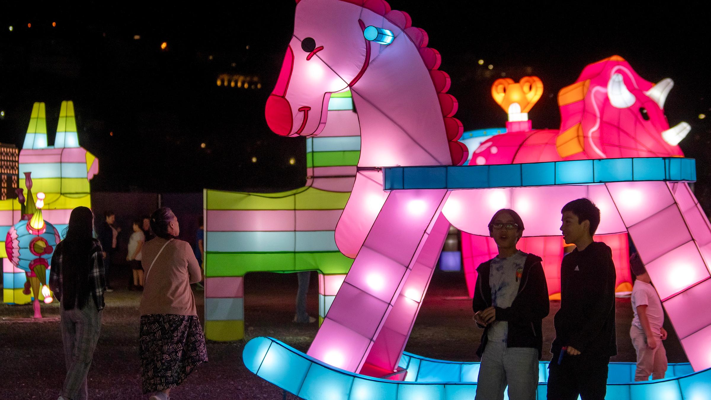 Auf einem Platz stehen weit voneinander entfernt verschiedene Lichtinstallationen von Tieren und Fabelwesen, daneben laufen einzelne Passanten. Die vorderste Installation zeigt ein pinkes Schaukelpferd.