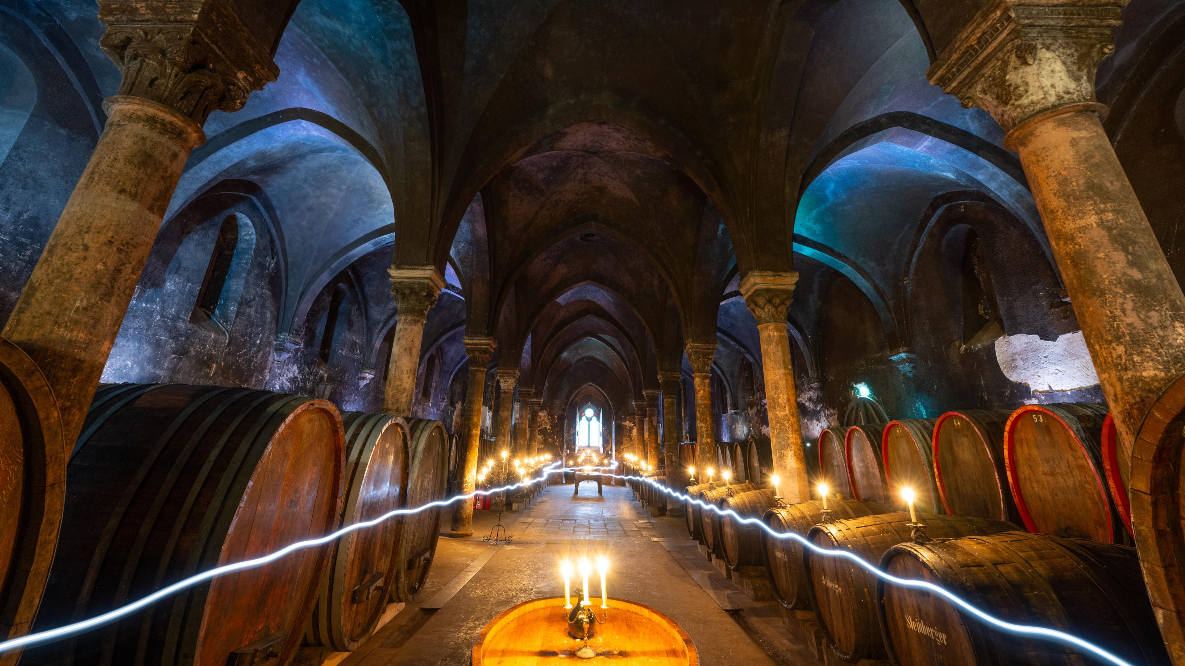 Blick durch die Mitte eines großen Kellergewölbes eines Klosters mit Weinfässern am Rand. Das Gewölbe ist mit Kerzen beleuchtet und ein Lichtstreifen erzeugt durch Handy-Light-Painting zieht sich links und rechts durch den Gang in der Mitte.