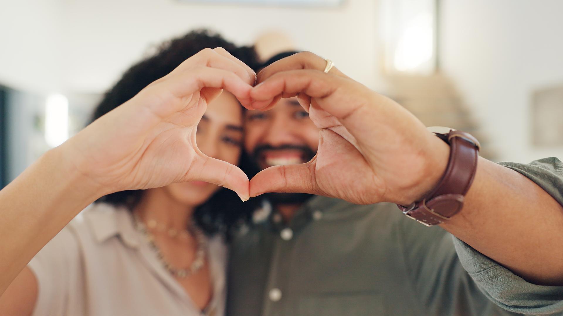 Typical für Beziehung und Liebe: Ein paar Bildet formt mit dem Händen ein Herz Richtung Kamera, im Hinergrund erkennt man ihre lächelnden Gesichter.