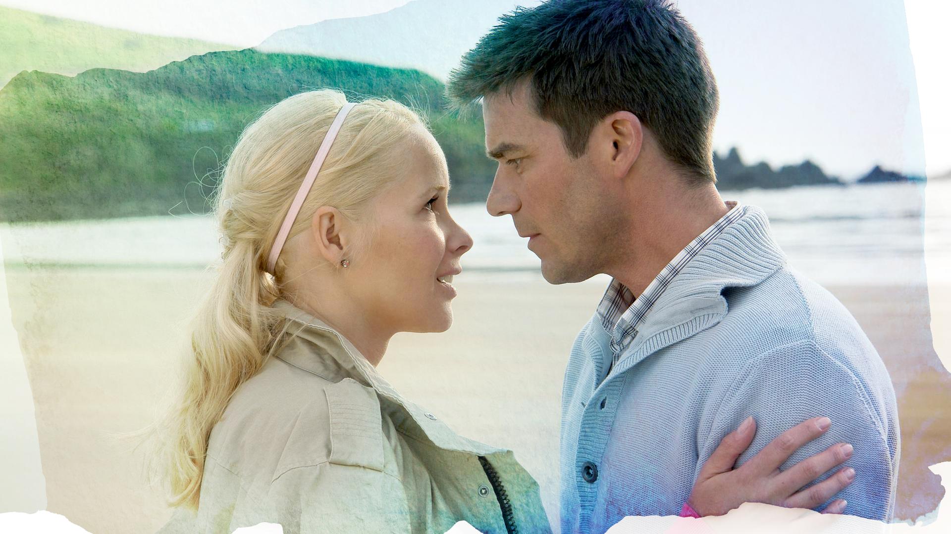 Rachel Wilson (Sylvia Leifheit, links) und Luke Folder (Thomas Scharff, rechts) stehen sich gegenüber und halten sich in den Armen. Ihre Gesichter sind sich sehr nah und sie schauen einander direkt in die Augen.