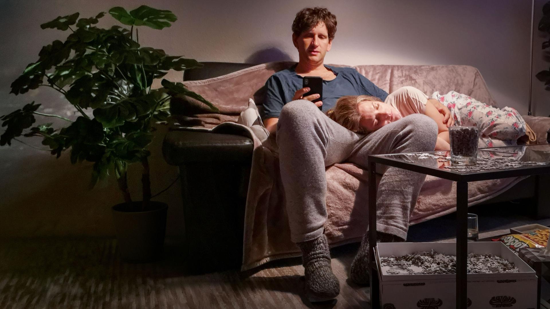 "Liebe. Jetzt! - Der Kuss":  Heiner (Sebastian Schwarz) sitzt mit seinem Handy auf dem Sofa. Seine Frau Meike (Marie Burchard) liegt schlafend auf seinem Schoß.