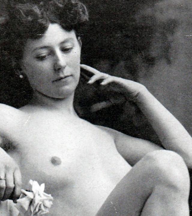 Das Bild zeigt eine schwarz-weiße Fotografie einer weiblichen Person, die nackt ist. Sie sitzt entspannt und hat eine nachdenkliche Haltung eingenommen, wobei sie mit ihrer rechten Hand an ihrem Gesicht spielt und mit der linken Hand eine Blume hält. Ihr Haar ist zu kleinen Locken frisiert. Der Hintergrund ist unscharf, sicher aber ein neutraler Farbton, um die Person hervorzuheben. Die Pose und der Ausdruck der Person vermitteln eine gewisse Intimität und nachdenkliche Stimmung.   Diese Art der Fotografie spiegelt den sozialen Wandel und das neu entdeckte Körperbewusstsein des späten 19. Jahrhunderts wider, wie im Kontext des mitgelieferten Textes erwähnt.