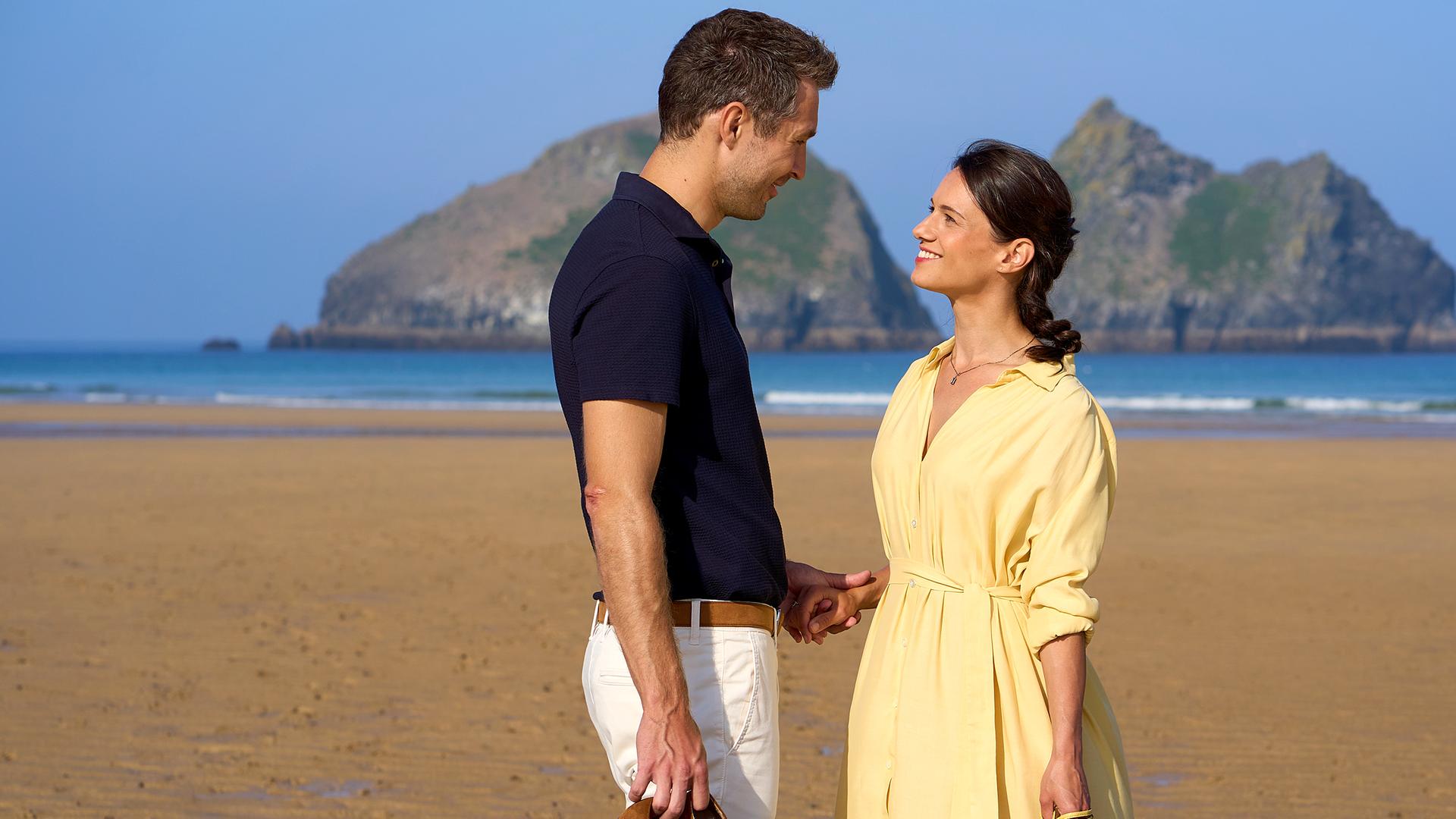Carl Coburn (Jan-Martin Müller) steht am Strand, hält Mia Barlow (Leni Adams) an der Hand und sieht sie lächelnd an.