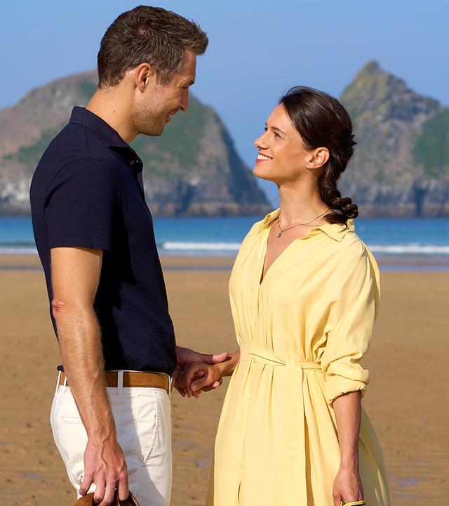Carl Coburn (Jan-Martin Müller) steht am Strand, hält Mia Barlow (Leni Adams) an der Hand und sieht sie lächelnd an.