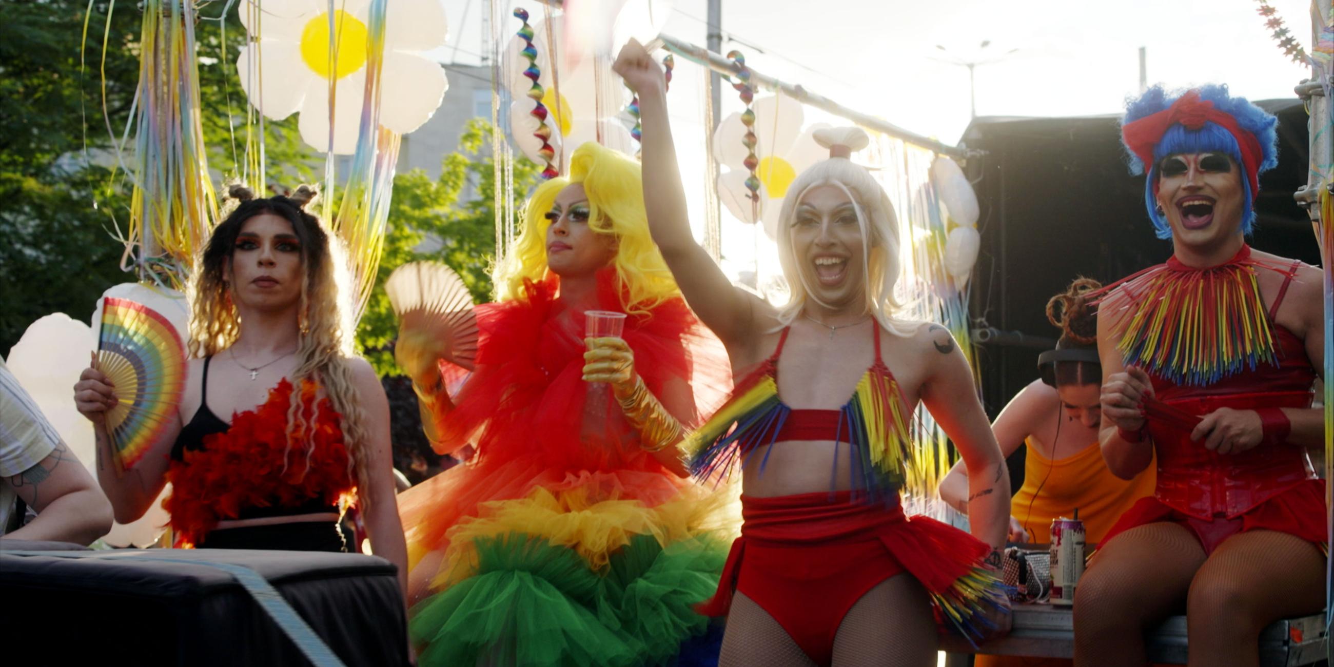 Vier Dragqueens feiern auf einem Festwagen die Pride Parade. Sie tragen bunte Perücken und Kostüme.