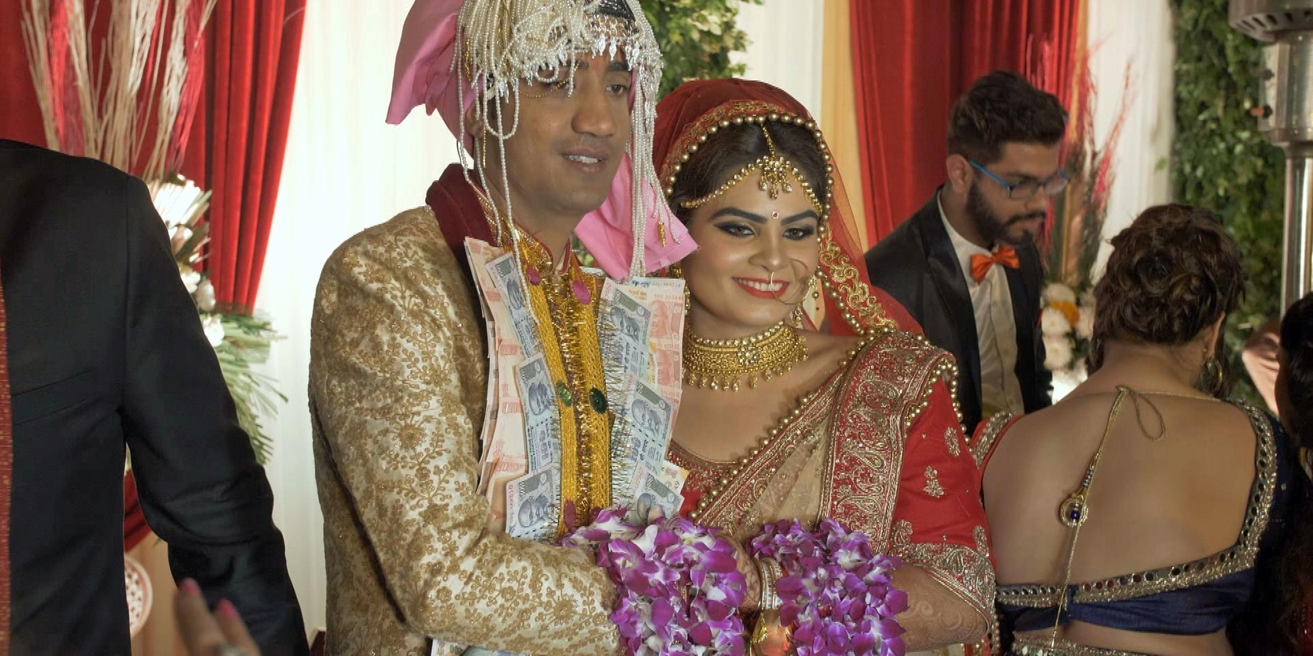 "Liebe und Sex in Indien (AT)": Zentral ein prächtig gekleidetes und geschmücktes Brautpaar inmitten einer privaten Hochzeitsgesellschaft. An seinen Revers sind unzählige Banknoten angeheftet, sie stehen eng nebeneinander, halten sich an den Händen, über ihre Unterarme sind lila Blumengirlanden drapiert.
