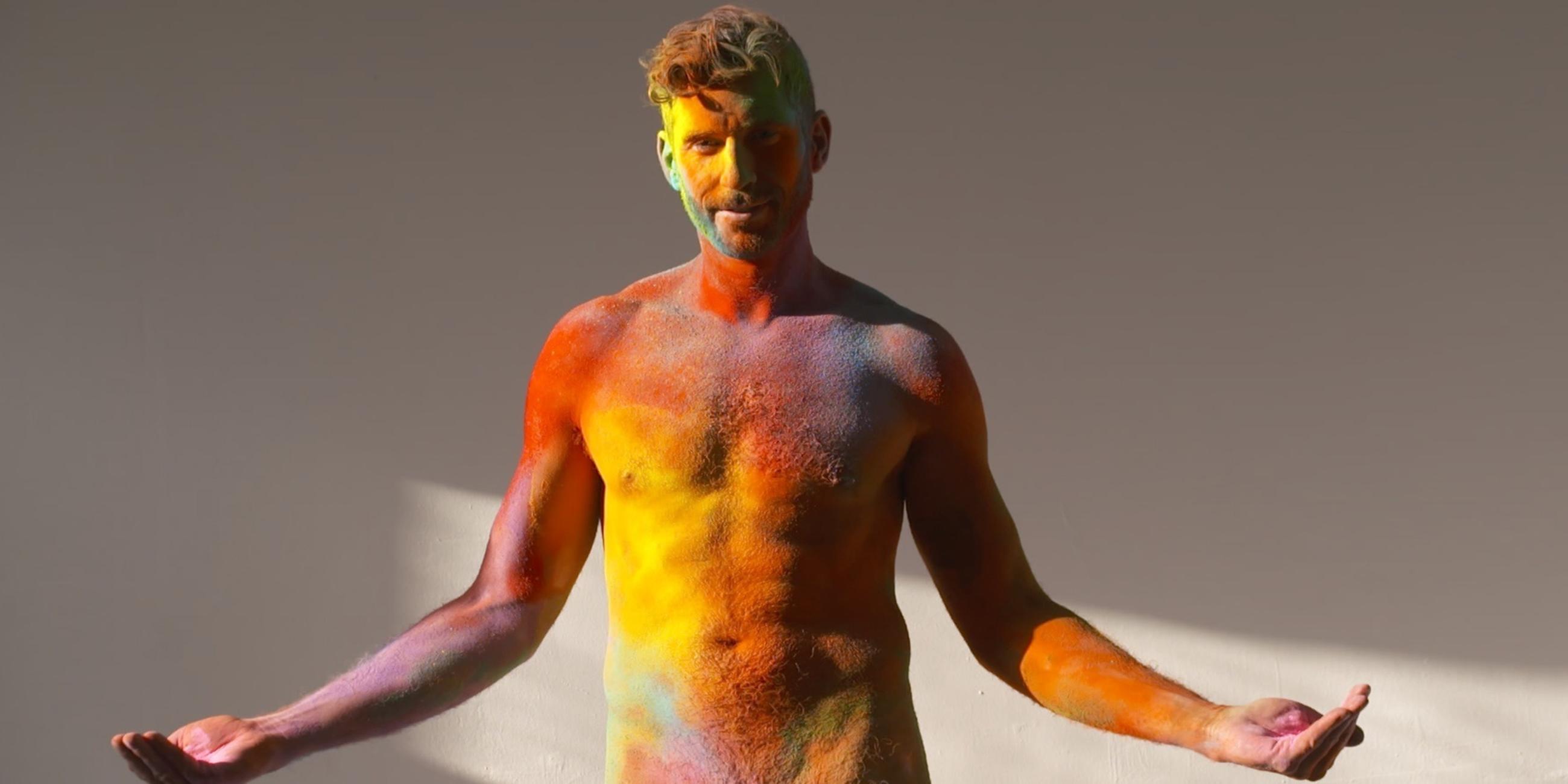 Ein nackter Mann streckt die Arme von sich und schaut in die Kamera. Sein Körper ist mit verschiedenen Farben bemalt.