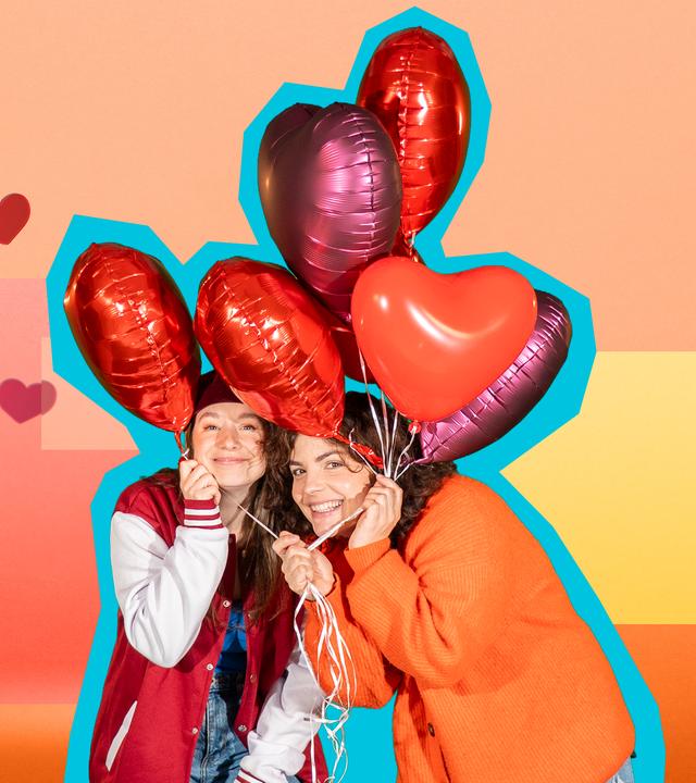 Phela und Senta halten mehrere Herzluftballons in den Händen und lächeln in die Kamera. Der Hintergrund ist gelb-orange-rot.