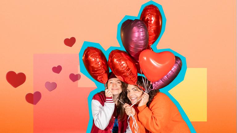 Phela und Senta halten mehrere Herzluftballons in den Händen und lächeln in die Kamera. Der Hintergrund ist gelb-orange-rot.