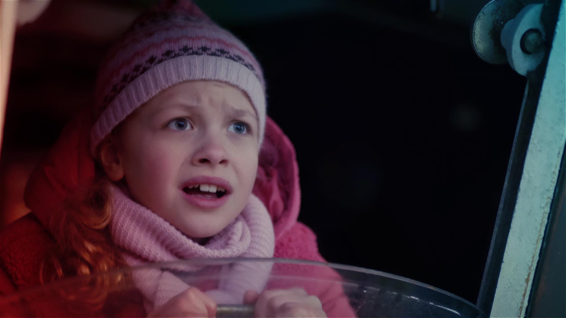 Ein Mädchen im Grundschulalter, das rosa Winterkleider trägt, schaut verängstigt aus einem Fenster heraus, das zu einem Boot gehört.
