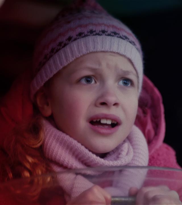 Ein Mädchen im Grundschulalter, das rosa Winterkleider trägt, schaut verängstigt aus einem Fenster heraus, das zu einem Boot gehört.