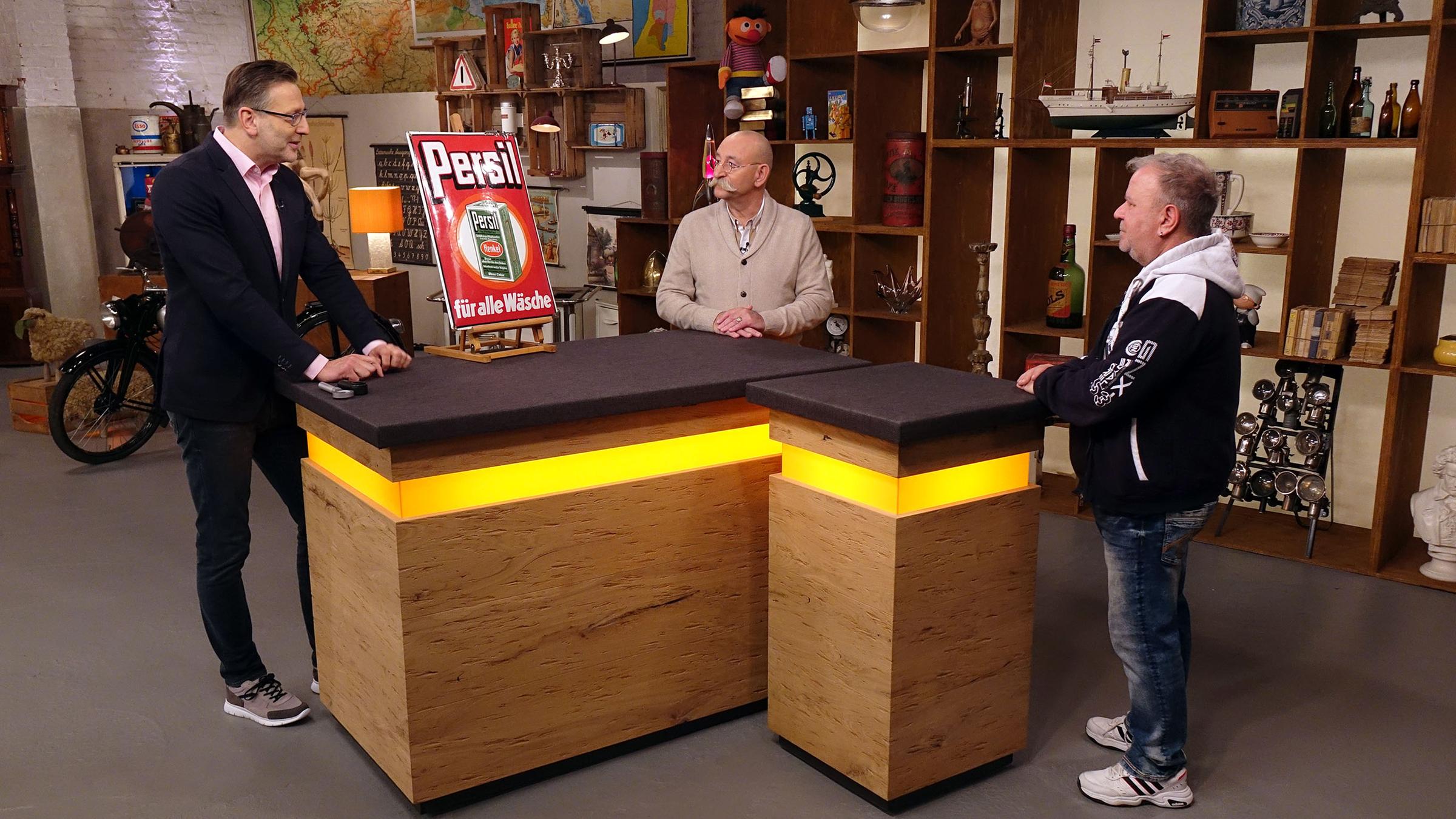 Experte Detlev Kümmel begutachtet ein Emailleschild von Persil. Neben ihm am Expertisentisch stehen Horst Lichter und der Verkäufer