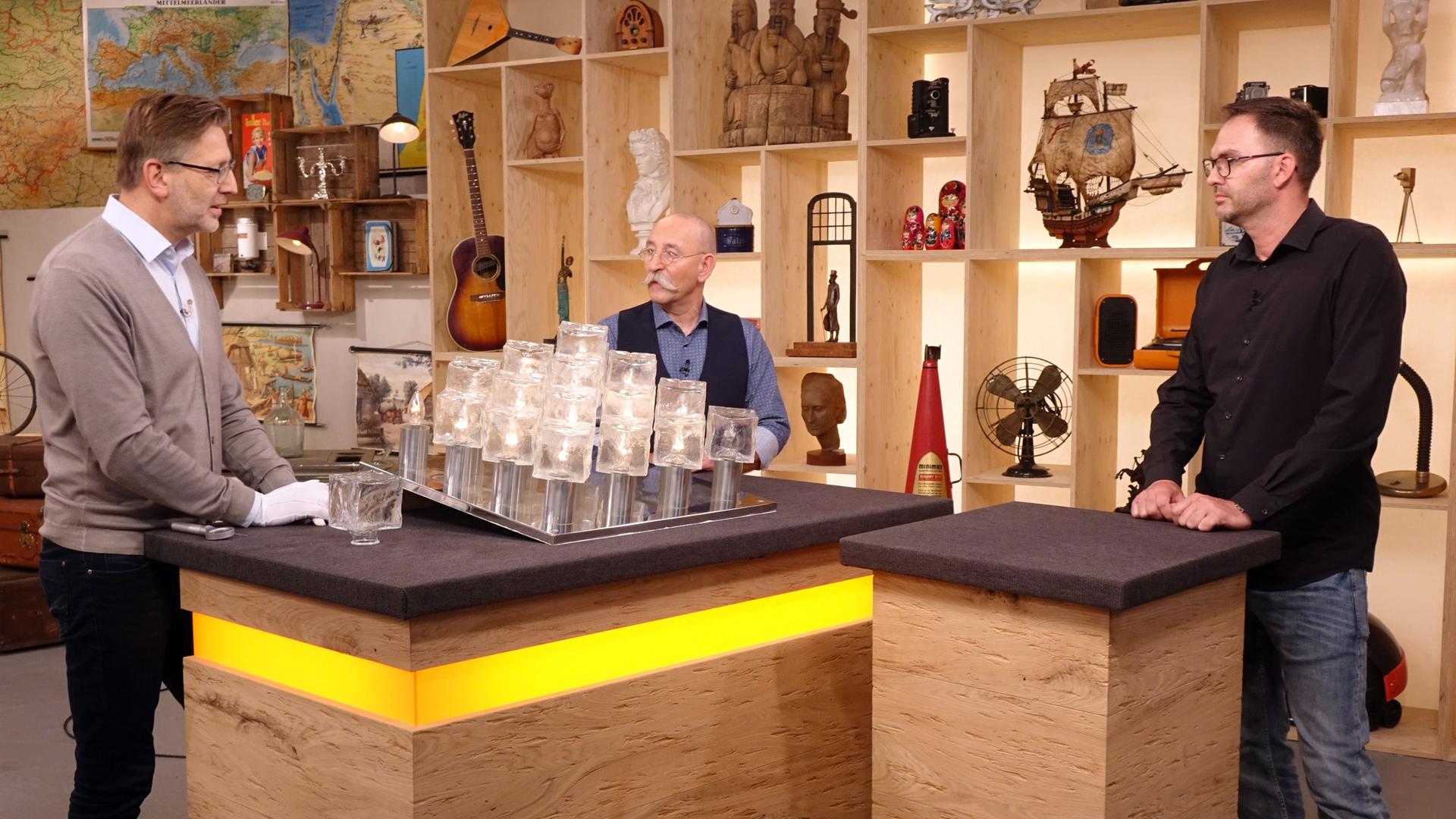 Experte Detlev Kümmel begutachtet eine "Ice Cube"-Deckenleuchte. Mit ihm am Expertenpult stehen Horst Lichter und der Verkäufer der Lampe.