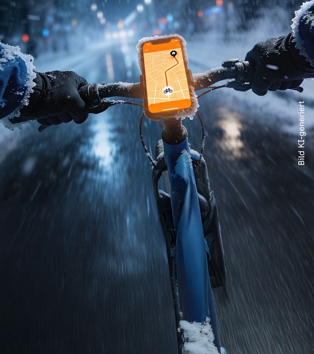 Ein Fahrradlenker, an dem ein Handy festgemacht ist, das eine Route anzeigt. Zwei Hände am Rand des Fahrradlenkers. Im Hintergrund ist die Straße zu erkennen.