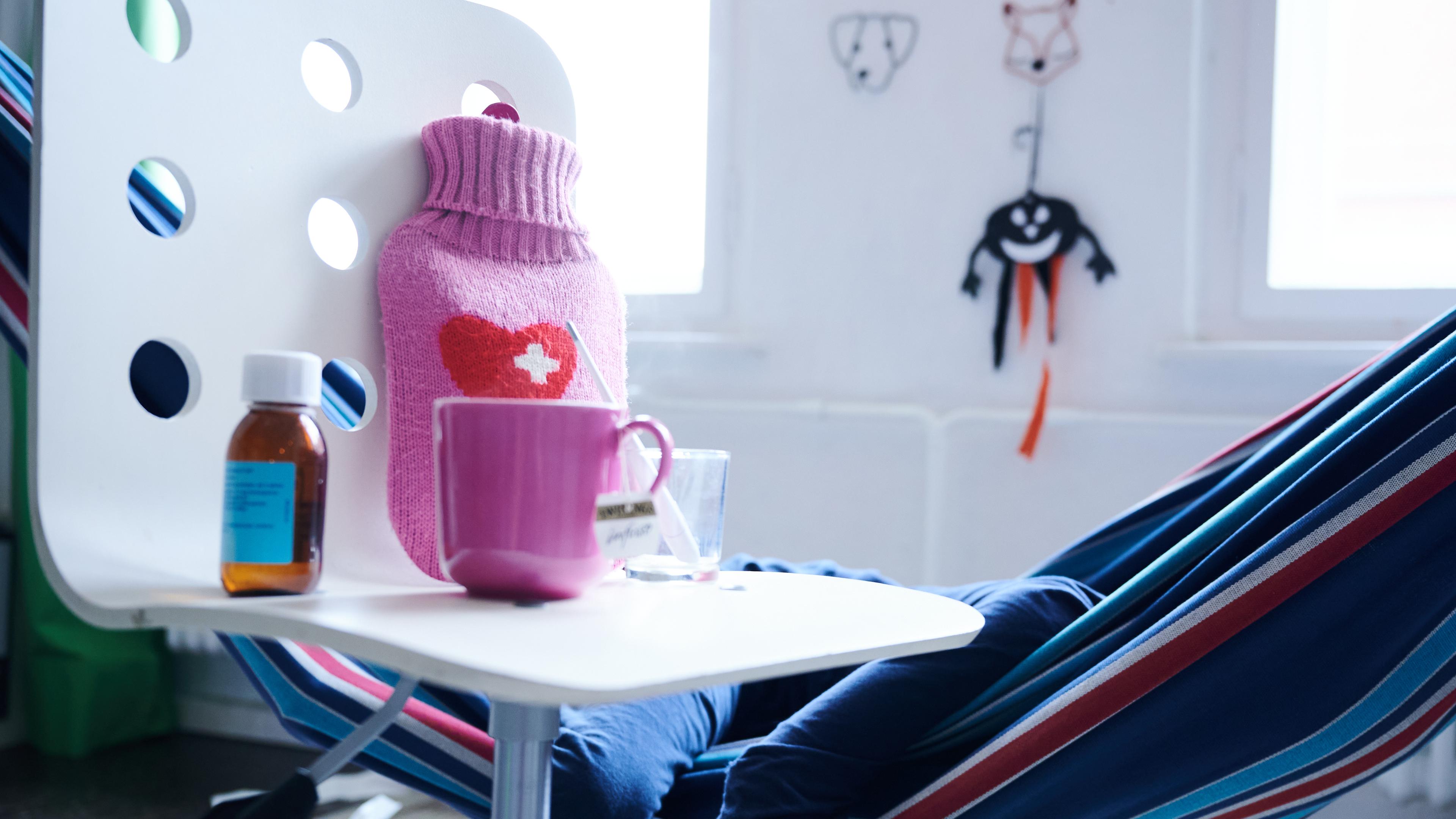  Fieberthermometer, Hustensaft, Wärmflasche und eine Tasse Tee stehen auf einem Stuhl, während ein Kind im Hintergrund in einer Hängematte liegt 