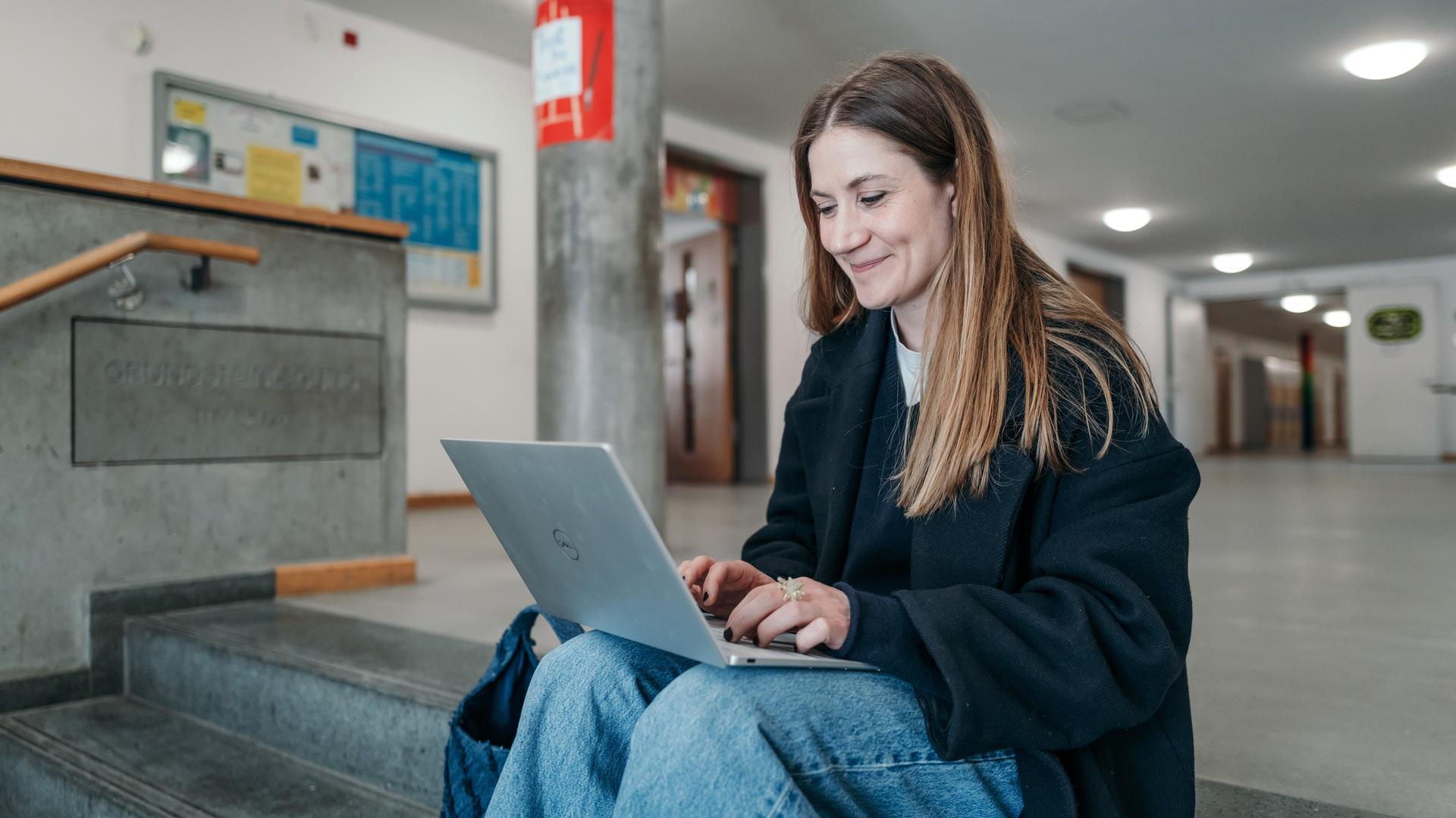 Charlotte sitzt auf einer Treppe, hat ihren Laptop auf ihre Beine vor sich gestellt und lächelt in den Laptop. 