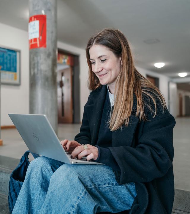 Charlotte sitzt auf einer Treppe, hat ihren Laptop auf ihre Beine vor sich gestellt und lächelt in den Laptop. 