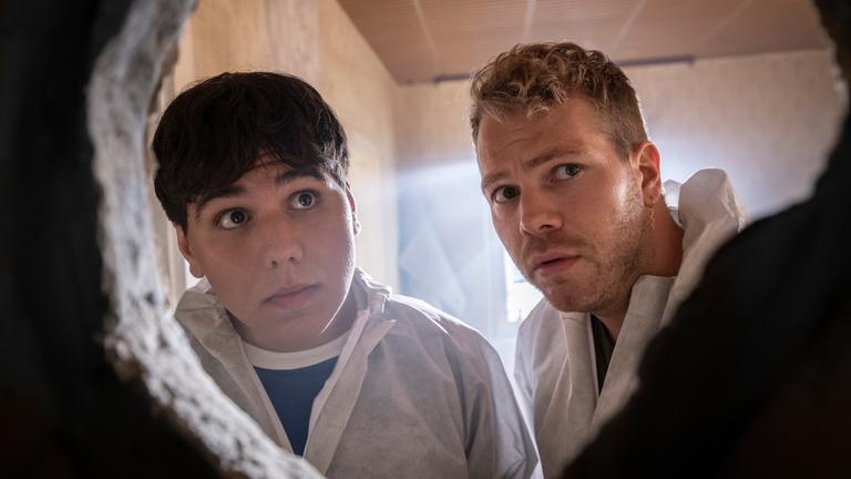 "Like a Loser": Ernst Hakke (Diyar Ilhan) und Julian Beil (Ben Münchow) schauen durch ein großes Loch in der Wand. Beide tragen weiße Schutzanzüge, Ernst blickt überrascht und leicht verwirrt, während Julian besorgt und angespannt wirkt. Im Hintergrund fällt ein Lichtstrahl durch ein Fenster in das Zimmer und hebt die staubige, alte Wand hervor.