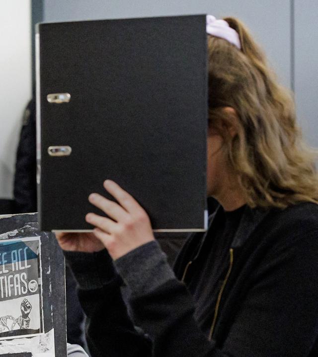 Zu sehen ist die Verurteilte Lina. E, die sich hinter einem Aktenordner versteckt; im Hintergrund ihre Verteidiger. 