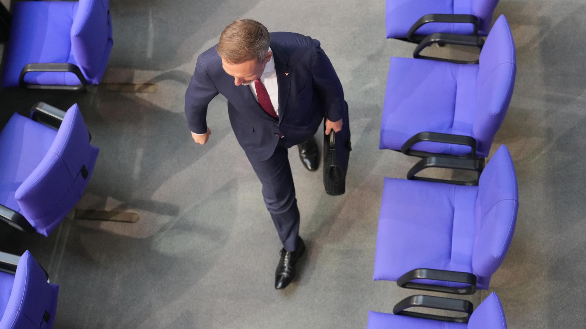 Draufsicht von oben auf einen Mann im Anzug, der mit einem Aktenkoffer in der Hand an einer leeren Stuhlreihe im Plenarsaal vorbeiläuft.