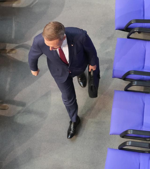 Draufsicht von oben auf einen Mann im Anzug, der mit einem Aktenkoffer in der Hand an einer leeren Stuhlreihe im Plenarsaal vorbeiläuft.