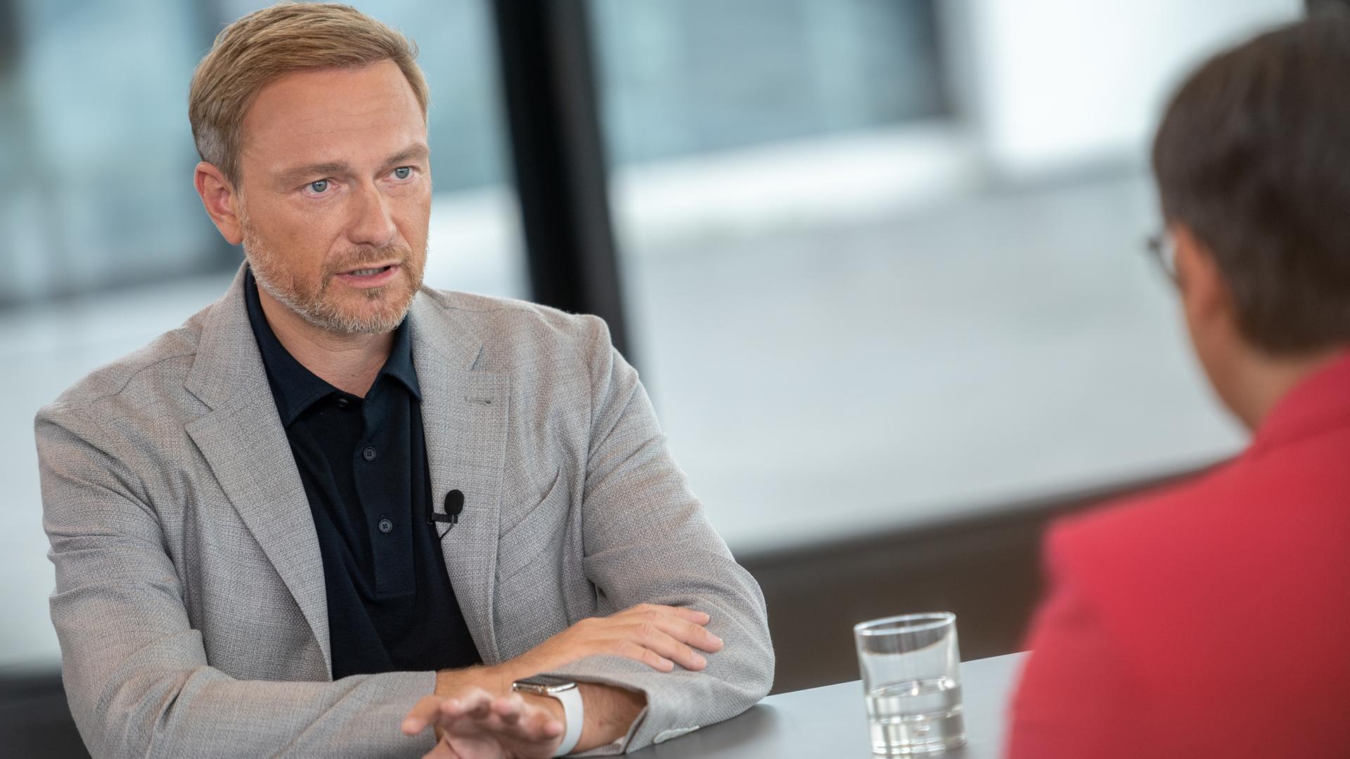 Christian Lindner sitzt an Tisch mit Glas Wasser. Unscharf im Vordergrund: Frau Banerjee mit rotem Blazer.