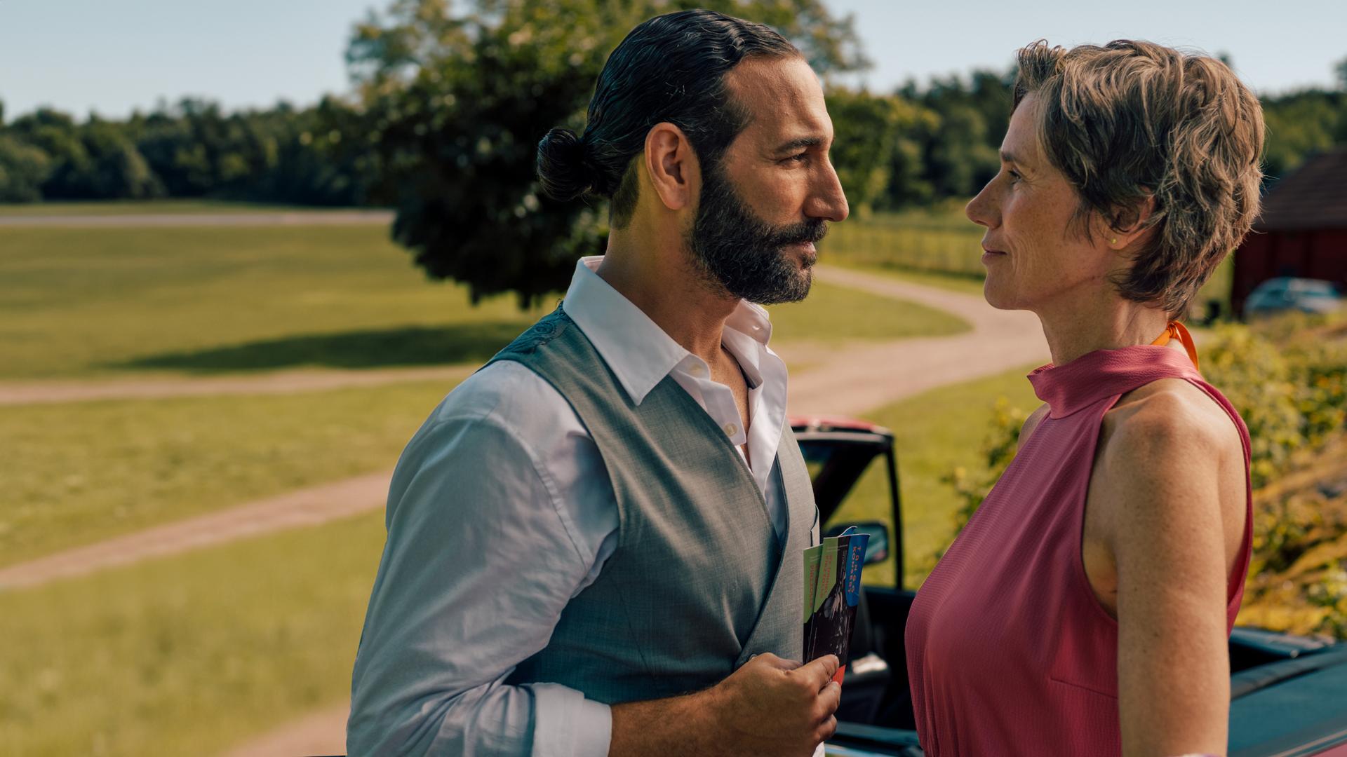 "Inga Lindström: Rezept für die Liebe": Frederika (Julia Bremermann) und Kian (Massimo Sinato) stehen im Freien dicht beieinander und sehen sich an. Im Hintergrund liegt eine sich schlängelnde Schotterstraße, umgeben von Bäumen und Wiese. Direkt hinter dem Paar steht ein rotes Cabrio mit offenem Dach.