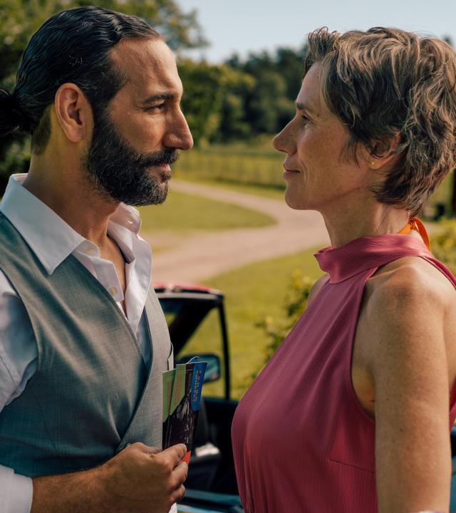 "Inga Lindström: Rezept für die Liebe": Frederika (Julia Bremermann) und Kian (Massimo Sinato) stehen im Freien dicht beieinander und sehen sich an. Im Hintergrund liegt eine sich schlängelnde Schotterstraße, umgeben von Bäumen und Wiese. Direkt hinter dem Paar steht ein rotes Cabrio mit offenem Dach.