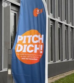 Blau-orange Fahne mit dem Schriftzug "Pitch dich!" und dem ZDF-Logo vor einer grauen Gebäudefassade.