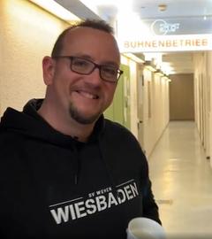 Mann mit SV Wehen Wiesbaden Pullover lächelt in die Kamera: im Hintergrund Deckenschild im Flur "ZDF-Bühnenbetrieb"