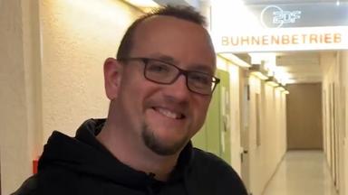 Mann mit SV Wehen Wiesbaden Pullover lächelt in die Kamera: im Hintergrund Deckenschild im Flur "ZDF-Bühnenbetrieb"