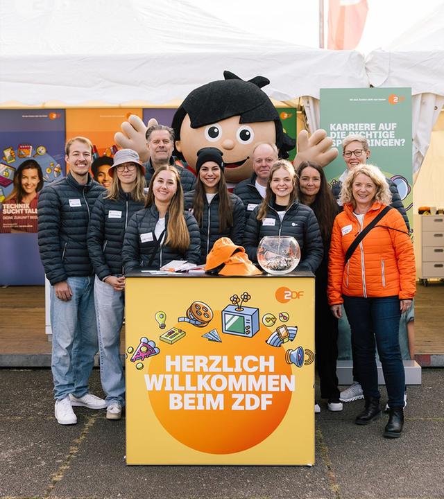 Gruppenfoto des ZDF-Teams auf der BIM: Zehn Teammitglieder und ein Mainzelmännchen versammeln sich hinter einem gelben Messetresen. Informationsmaterialien, eine Lostrommel und Plakate zum Thema Karriere beim ZDF kennzeichnen den Stand als Anlaufstelle für Ausbildungs- und Berufsinformationen