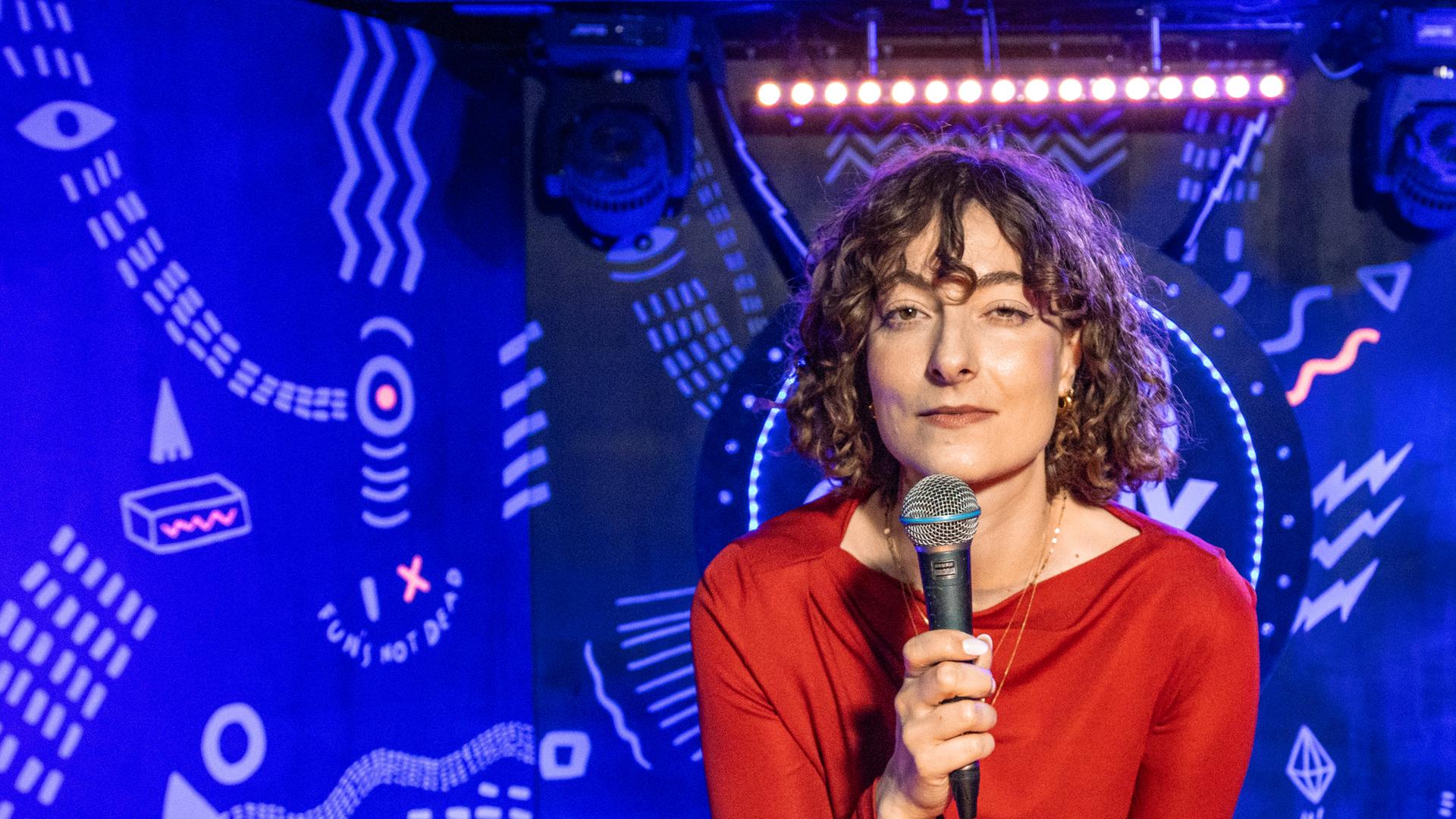 Lisa Frischemeier steht in der Mitte des Bildes und hat ein Mikrofon in der Hand. Der Hintergrund ist blau beleuchtet.
